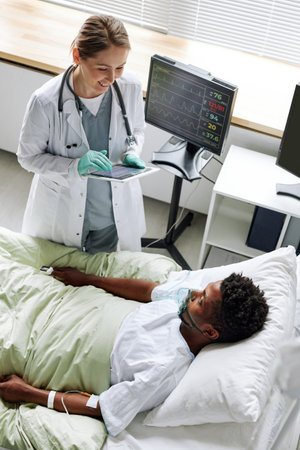 Vertical high angle view of cheerful young adult doctor greeting young Black man after waking up from comaの写真素材