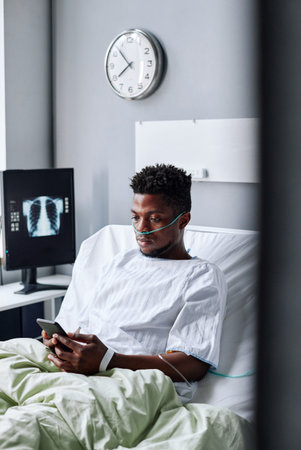 Young African American man spending days in hospital ward killing time surfing Internet on smartphoneの写真素材
