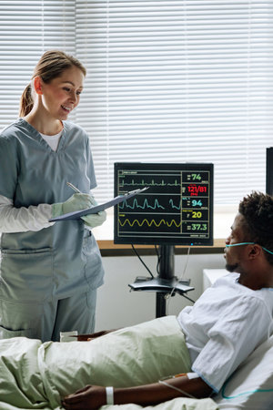 Vertical shot of cheerful Caucasian female doctor visiting young Black patient in hospital ward to check his health stateの写真素材