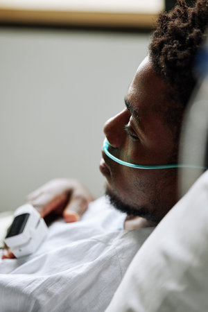 Vertical medium close-up of young Black man with nasal cannula and pulse oximeter on finger lying in hospital bed in wardの写真素材