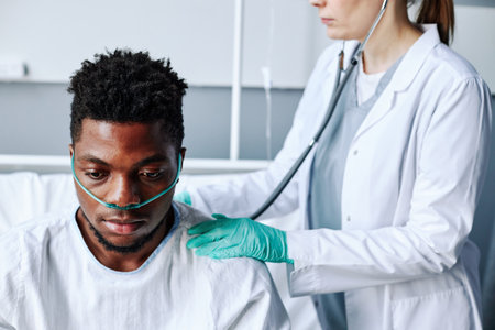 Selective focus shot of doctor working with young Black patient in hospital ward checking his health state using stethoscopeの写真素材