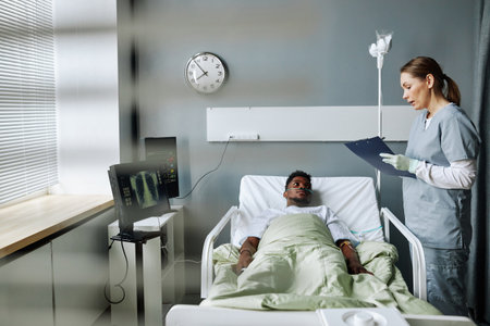 Caucasian female nurse holding clipboard with medical record standing in hospital ward asking questions to Black patientの写真素材