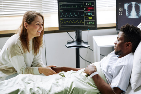 Cheerful young adult Caucasian woman visiting her Black boyfriend in hospital ward holding his hand and smilingの写真素材
