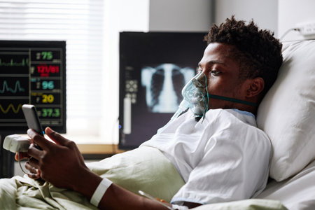 Horizontal shot of young Black man spending days in hospital ward scrolling news feed in Internet on smartphoneの写真素材