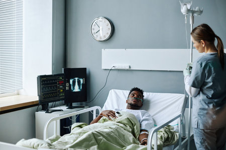 Young African American man with nasal cannula lying in hospital bed in ward waiting for nurse setting IV lineの写真素材