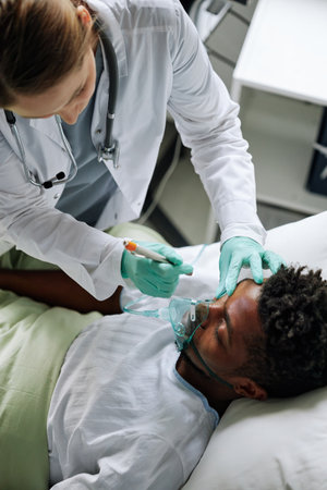 High angle of doctor checking health state of young Black man in coma using flashlight to see pupils movementの写真素材