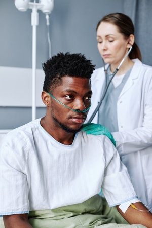 Vertical selective focus shot of young adult Caucasian doctor listening to internal sounds of Black patients heart and lungs using stethoscopeの写真素材