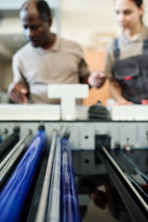 Selective focus shot of part of modern printer and two ethnically diverse coworkers standing by it,...