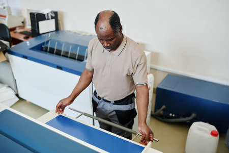 High angle view of African American man working in printing house adjusting blue cardstock paper on printer trayの写真素材