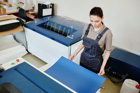 High angle view of young Caucasian worker standing in front of printing machine holding sheet of...