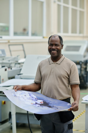 Vertical medium long portrait of happy African American man working in printing house holding poster smiling at cameraの写真素材