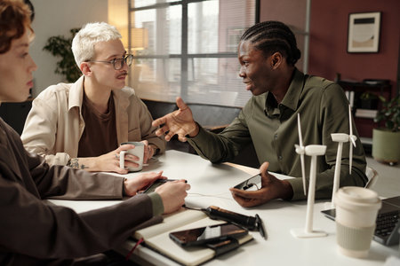 Group of three young ethnically diverse specialists sitting at office desk, discussing renewable...