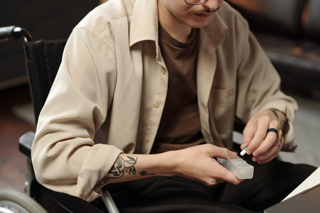 Medium section of young Caucasian man with disability sitting in wheelchair taking pill out of container at work in officeの写真素材