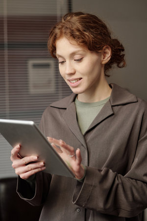 Vertical shot of young Caucasian woman with curly hair using digital tablet at work in modern office