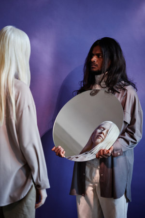 Vertical studio shot of handsome long-haired Indian man wearing casual clothes holding mirror...