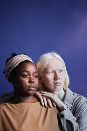 Vertical studio shot of young Black and Caucasian women standing against blue wall background looking awayの写真素材