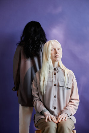 Vertical studio portrait of androgynous young woman with albinism sitting on chair looking away, unrecognizable man with long black hair standing behind herの写真素材