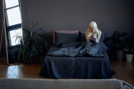 Horizontal high angle view shot of young Caucasian woman with albinism sitting on bed in early morning surfing Internet on smartphoneの写真素材