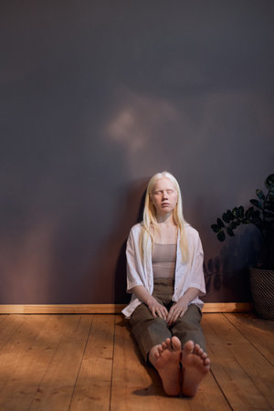 Vertical shot of beautiful young woman with albinism wearing casual outfit sitting on floor with eyes closedの写真素材