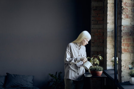 Horizontal shot of young woman with albinism wearing fashionable casual clothes watering houseplants in loft bedroom at homeの写真素材