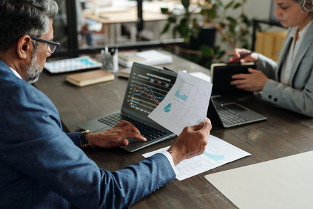 Middle aged Caucasian man analyzing financial charts on laptop while holding printed graphs, sitting...