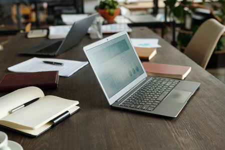 Top view showing open laptop displaying business analytics on wooden desk surrounded by notebooks,...