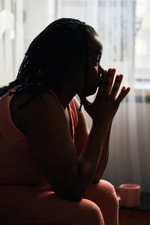 Vertical back light side view shot of young Black woman sitting in bedroom thinking about something