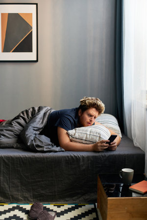 Vertical shot of young Caucasian man waking up in morning in his bedroom watching something on smartphoneの写真素材