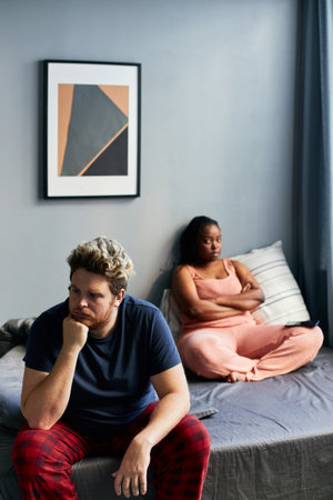 Vertical high angle shot young Caucasian man and his Black girlfriend sitting on bed in morning...