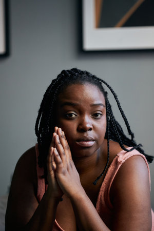 Vertical medium close-up shot of beautiful young Black woman sitting in bedroom at home looking at...