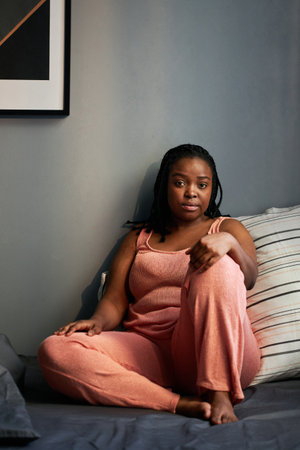 Vertical shot of beautiful young Black woman wearing stylish pyjamas sitting on bed in morning...