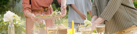 Woman serving platter of appetizers while another woman arranging table outdoors, teenager standing...