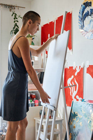 Vertical medium long shot of young woman placing canvas onto easel to start painting something