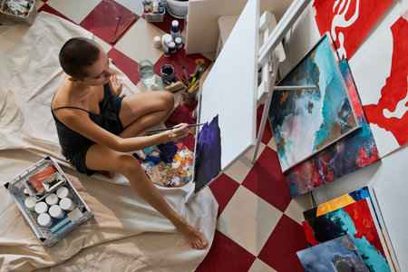 Horizontal high angle view shot of young professional artist sitting on floor in studio painting abstract picture on canvasの写真素材