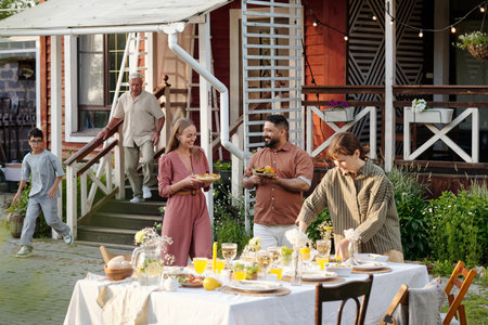 Group of multiethnic adults and one teenage boy gathering outdoors, smiling and preparing food around table set for meal, senior Caucasian man walking down steps in backgroundの写真素材