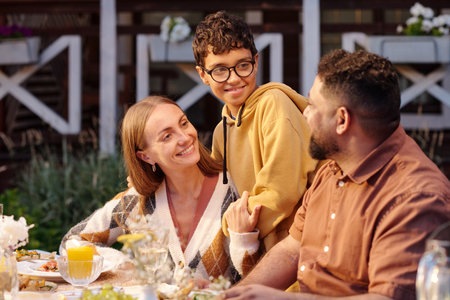 Caucasian woman, Middle Eastern man, and teenage boy interacting at outdoor table, teenage boy standing between adults smiling, adults looking at teenager, sharing family momentの写真素材