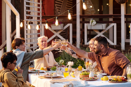 Multiethnic happy family sitting outdoors at table raising glasses in toast during festive meal, smiling and interacting togetherの写真素材