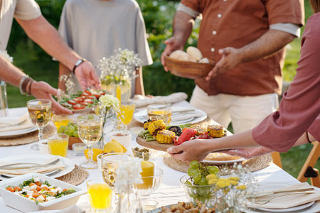 Multiethnic group of men and women serving grilled vegetables, salad, bread and drinks at outdoor table, hands reaching for food, enjoying summer gatheringの写真素材
