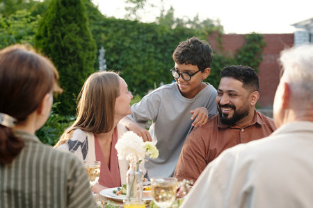 Multiethnic family of grandparents, young couple and their son gathering outdoors around table, smiling and interacting during shared mealの写真素材