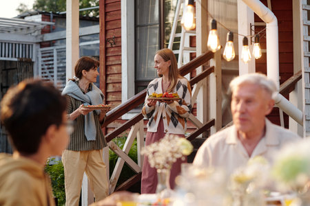 Two Caucasian women, one teenager and one middle aged, standing outdoors holding plates of food and smiling at each other while multiethnic group sitting at table in foregroundの写真素材