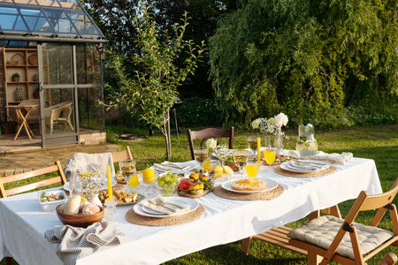 Outdoor table set for meal featuring plates with food, glasses of orange juice, bread basket, glass pitcher with water, arranged on tablecloth in garden near greenhouseの写真素材