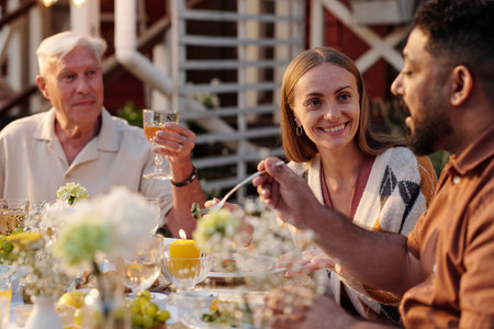 Caucasian senior man, Caucasian young adult woman, and Middle Eastern young adult man sitting outdoors sharing meal and conversation at table decorated with flowers and candlesの写真素材