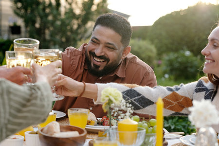 Middle Eastern man smiling while clinking glasses with diverse group of adults outdoors, enjoying...
