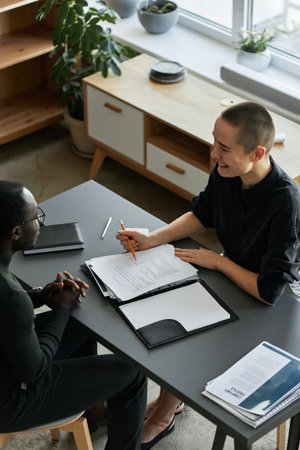 Vertical high angle view shot of cheerful young Caucasian woman sitting at desk in office having...