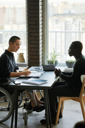 Young woman with disability sitting on wheelchair working on business strategy together with her Black male colleagueの写真素材