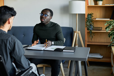Young African American HR manager interviewing Hispanic man with disability in wheelchair for vacancy, discussing work experienceの写真素材