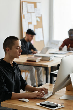 Vertical shot of stylish young Caucasian woman sitting at desk in modern office working on desktop...