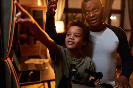 Modern Black father and his son spending evening together at home having fun watching stars using telescopeの写真素材