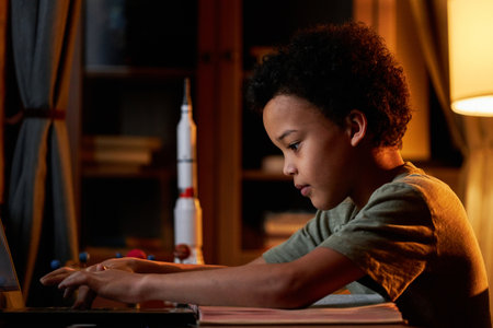 Side view portrait of gen Alpha Black boy sitting at desk in front of laptop in living room at home...