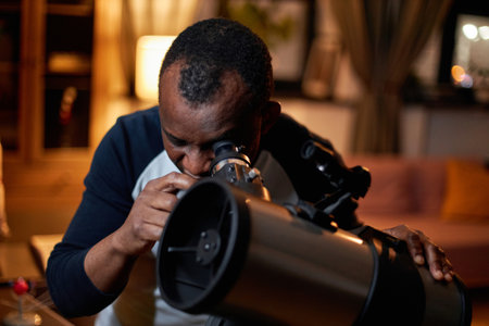 Modern mature African American man interested in astronomy enjoying watching stars and planets using telescope at homeの写真素材